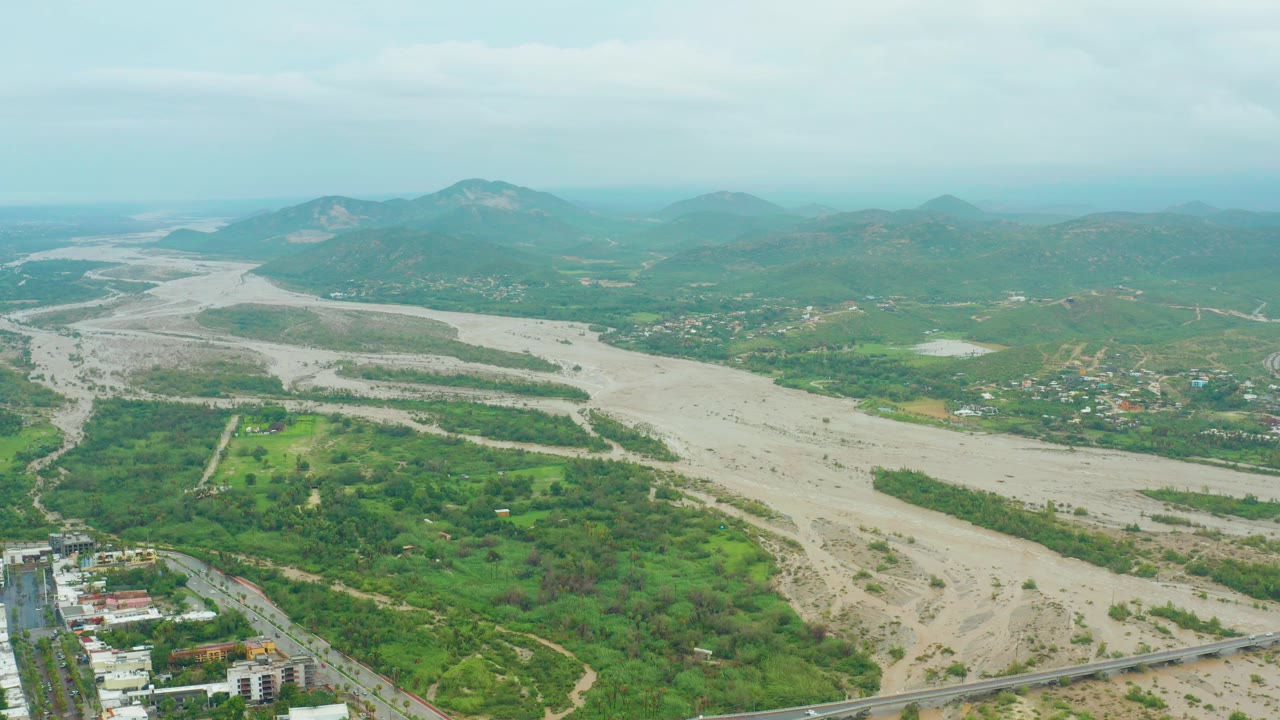 Aerial view in San Jose del Cabo, Baja California Sur, with a broad muddy river channel, green vegetation, scattered buildings, distant hills, and no visible people.