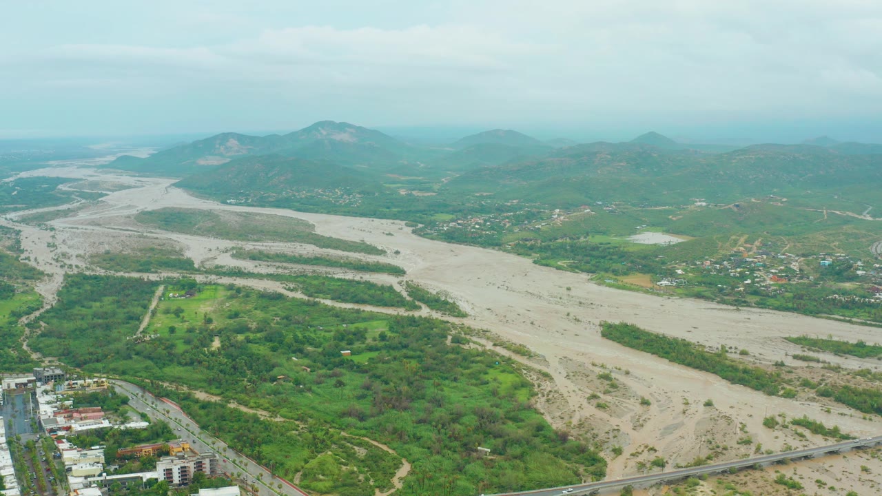 Aerial view over San Jose del Cabo with the muddy river running diagonally through green terrain, a small built area near the lower edge, hazy hills, and no visible people.