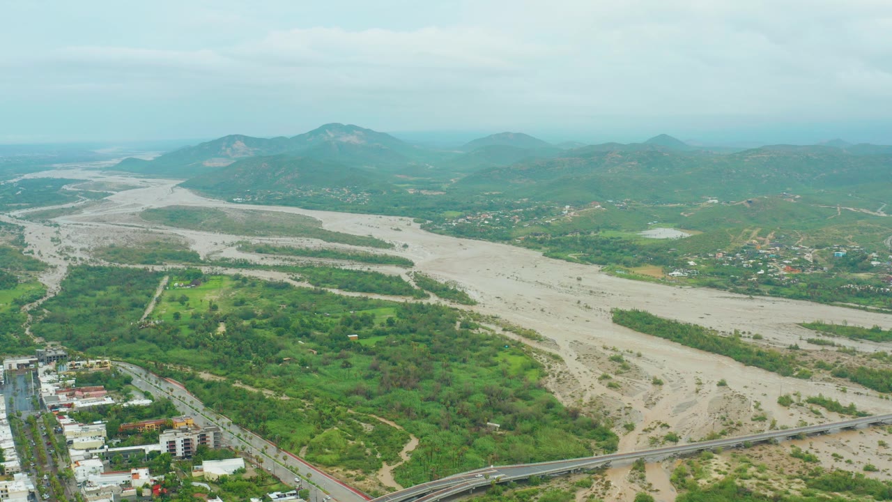 Aerial view in San Jose del Cabo, Baja California Sur with the pale river channel centered between green hills, a road and urban buildings in the lower frame, and no visible people.