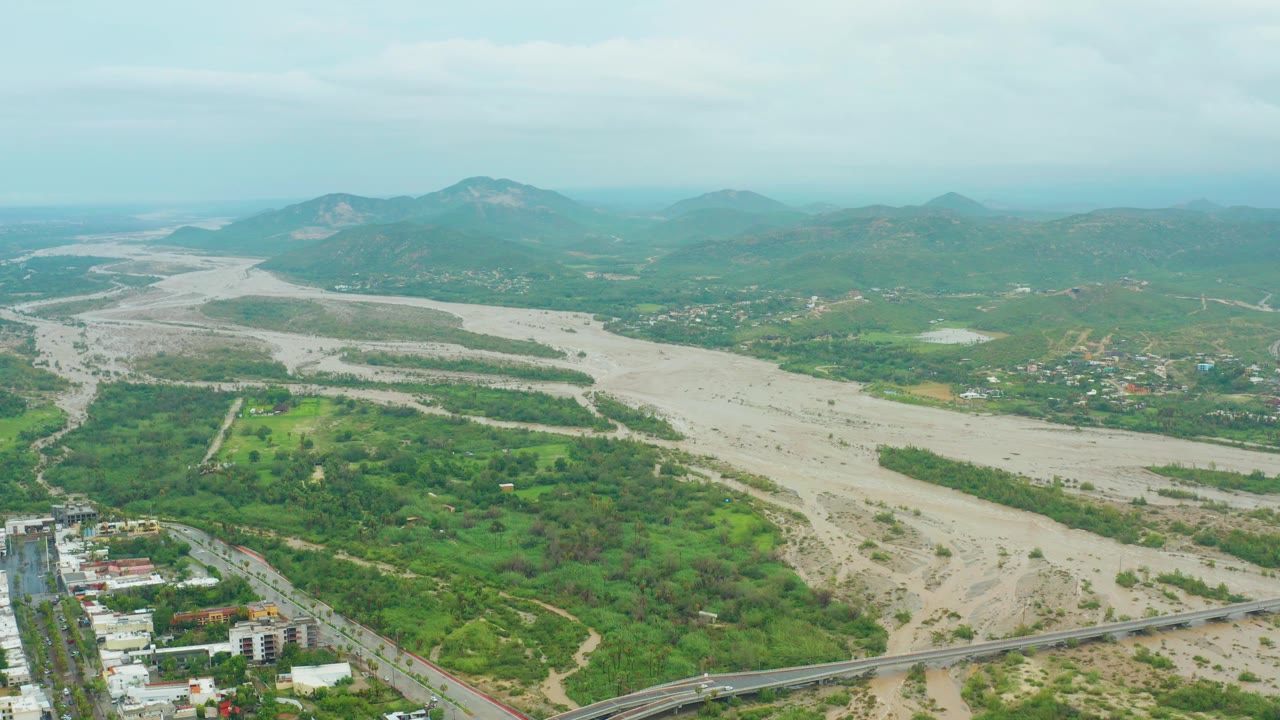 San Jose del Cabo aerial frame showing the riverbed stretching diagonally across green valley terrain, with clustered buildings and a road below and no visible people.