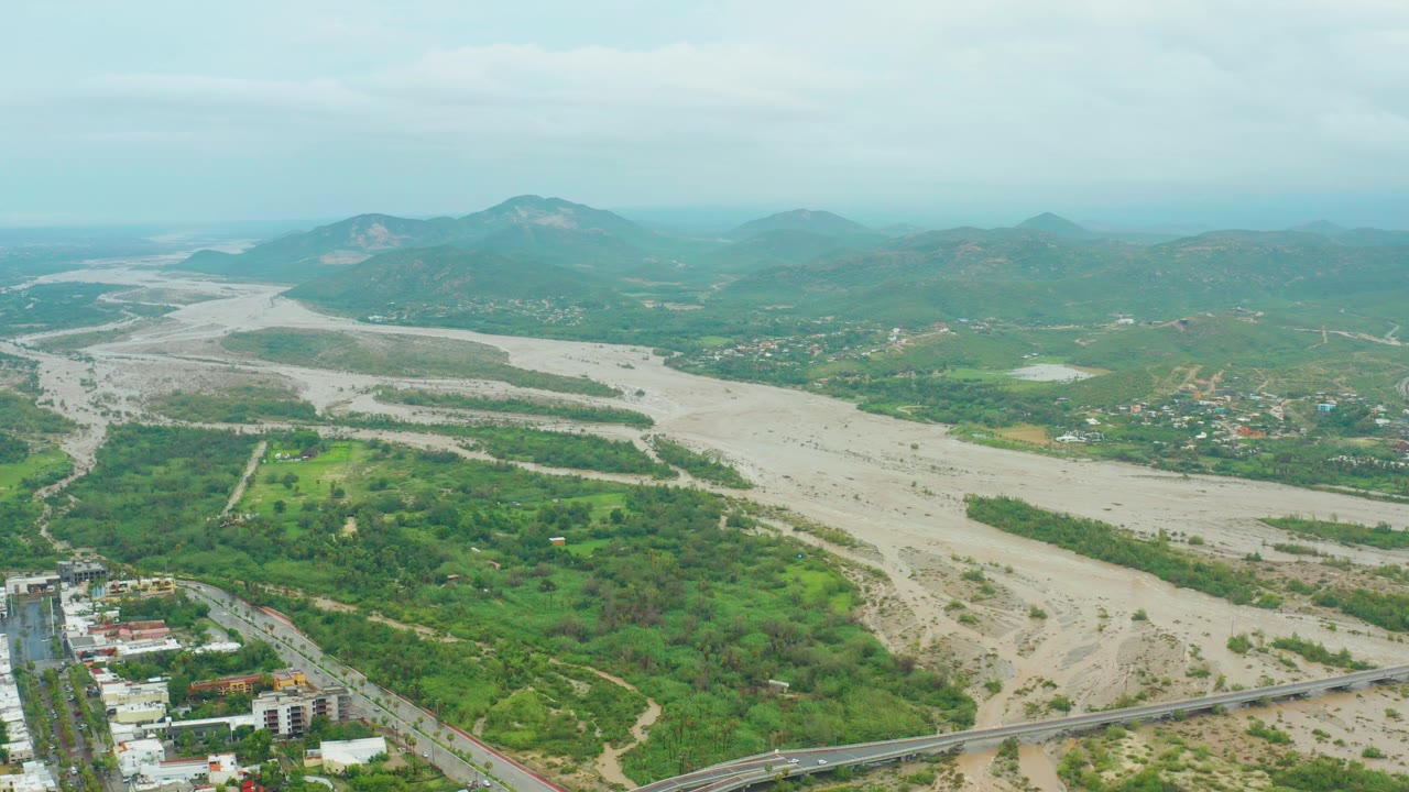 Mid-clip aerial view from San Jose del Cabo, Baja California Sur showing the pale riverbed widening through the valley, green hills around it, a bridge at the bottom, and no visible people.