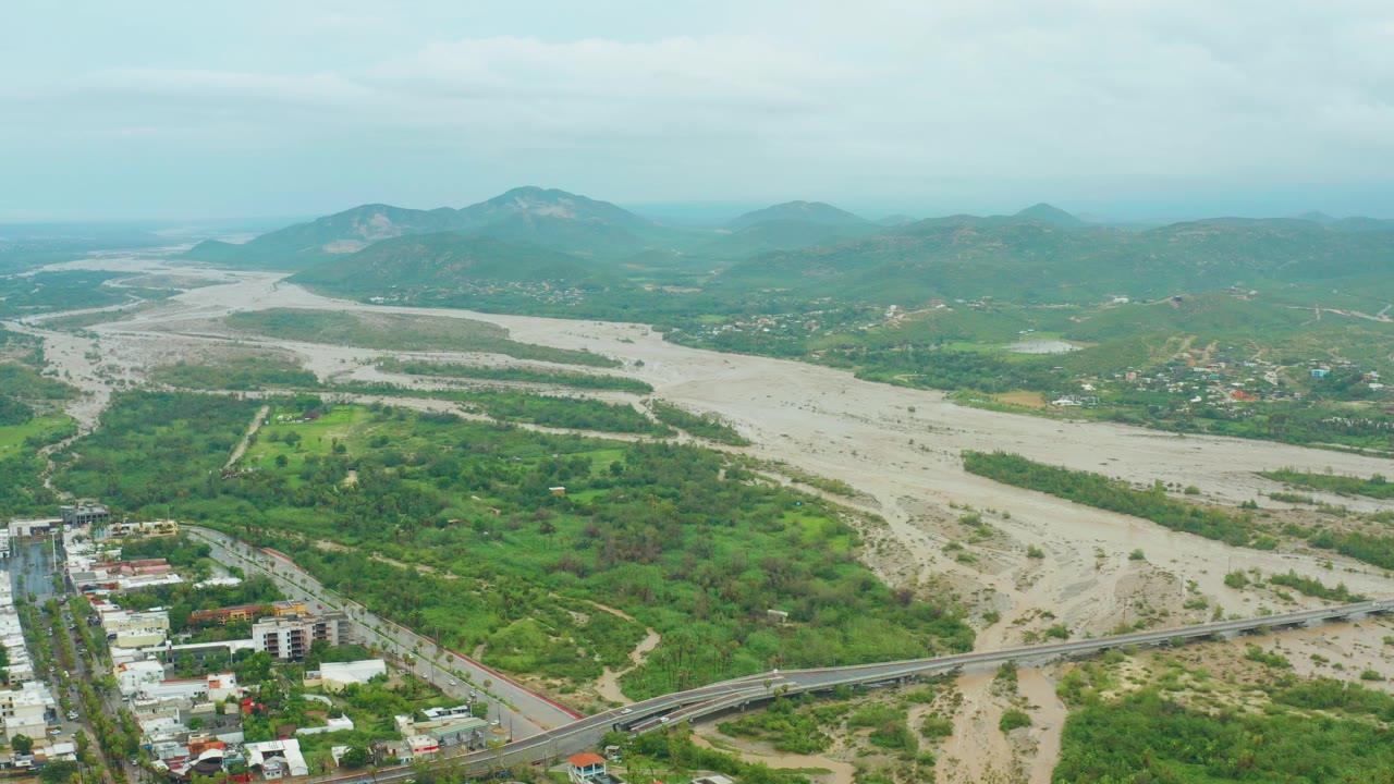 Aerial opening frame over San Jose del Cabo showing a wide pale riverbed, a bridge and road at the bottom edge, green hills beyond, distant mountains, overcast sky, and no visible people.