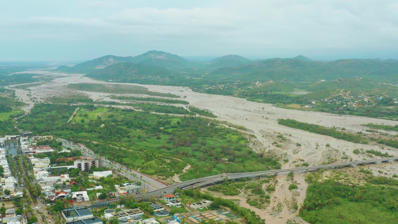 Wide aerial view of the muddy river channel with green vegetation, bridge, buildings, and mountains.