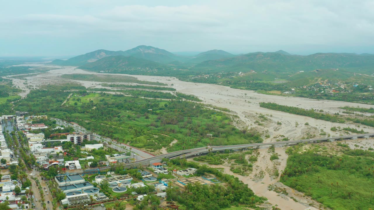 Wide aerial view in San Jose del Cabo, Baja California Sur, showing a muddy riverbed, bridge crossing, green vegetation, foreground buildings, distant hills, and no visible people.
