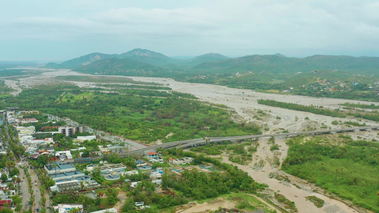 Opening aerial frame over San Jose del Cabo BCS Mexico showing a pale riverbed with muddy water channels, a bridge, green vegetation, low buildings, distant hills, overcast sky, and no visible people.