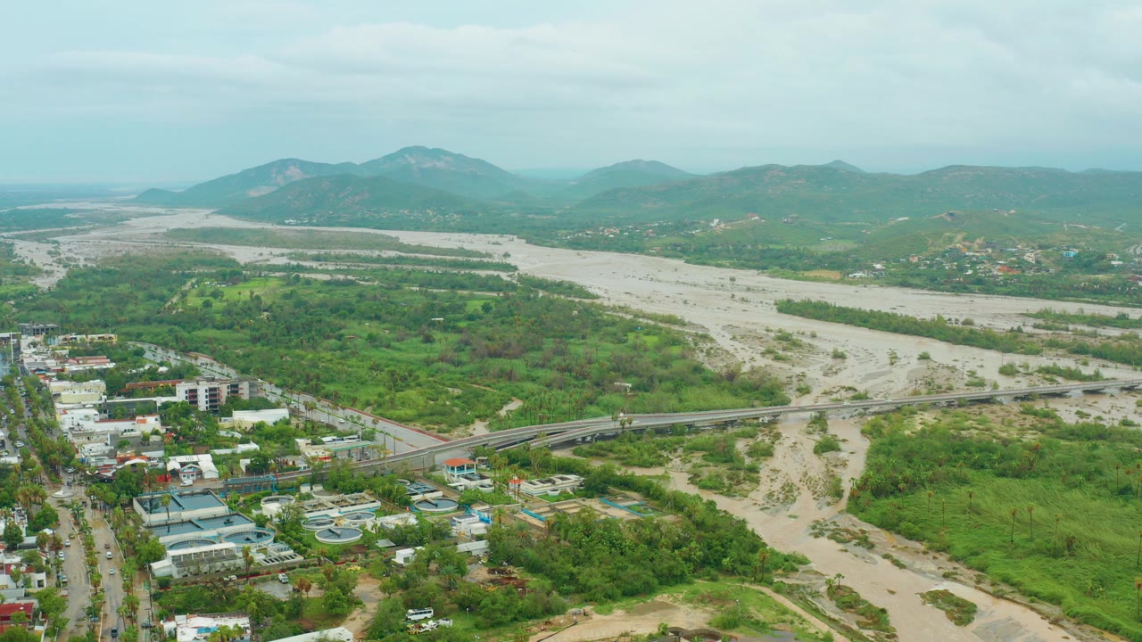 Second frame holds a high wide view in San Jose del Cabo BCS Mexico, with the bridge crossing the pale riverbed, clusters of buildings at lower left, green terrain across the valley, hazy mountains, and no visible people.