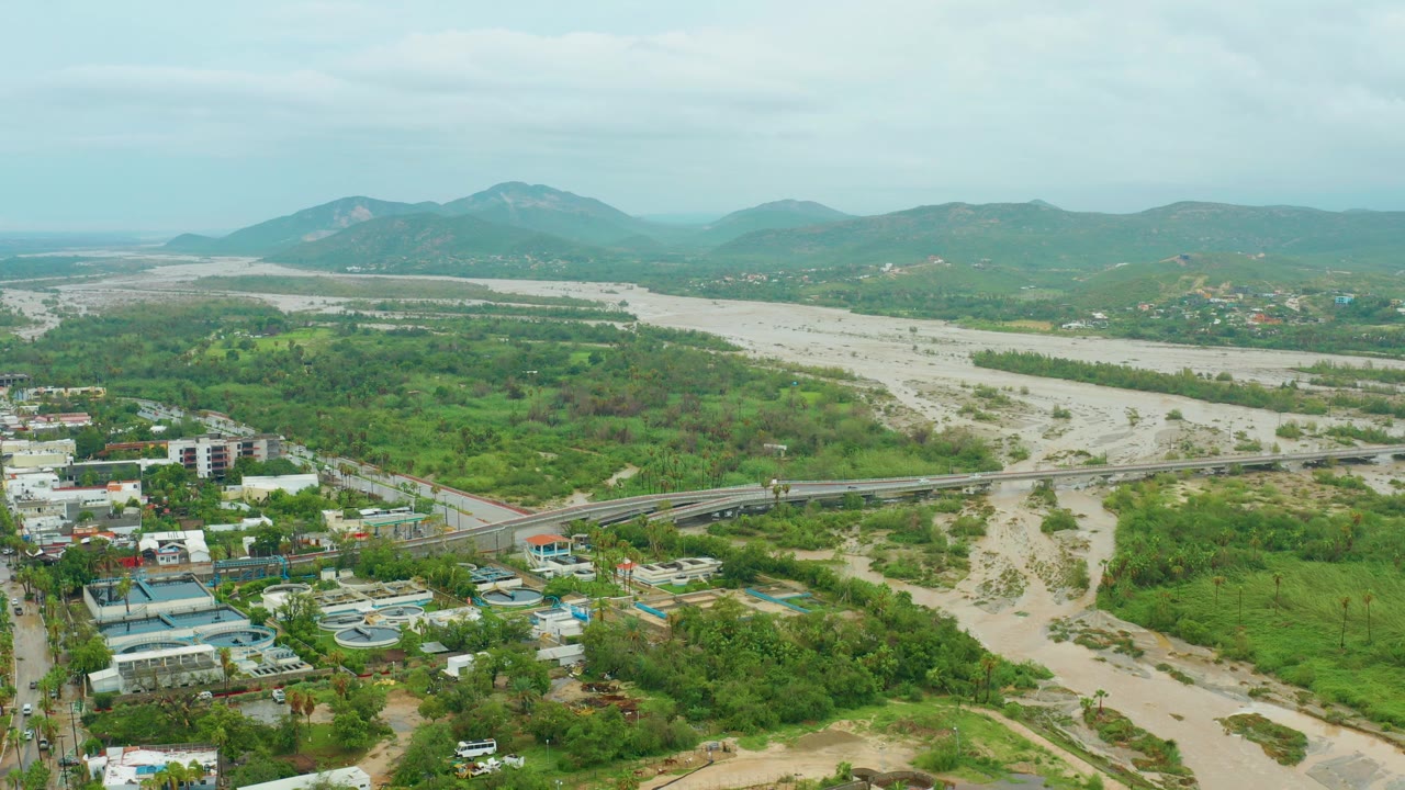 Aerial frame from San Jose del Cabo showing the broad light-colored riverbed, a bridge crossing near the lower center, roadside buildings to the left, dense green patches, hazy mountains, and no visible people.