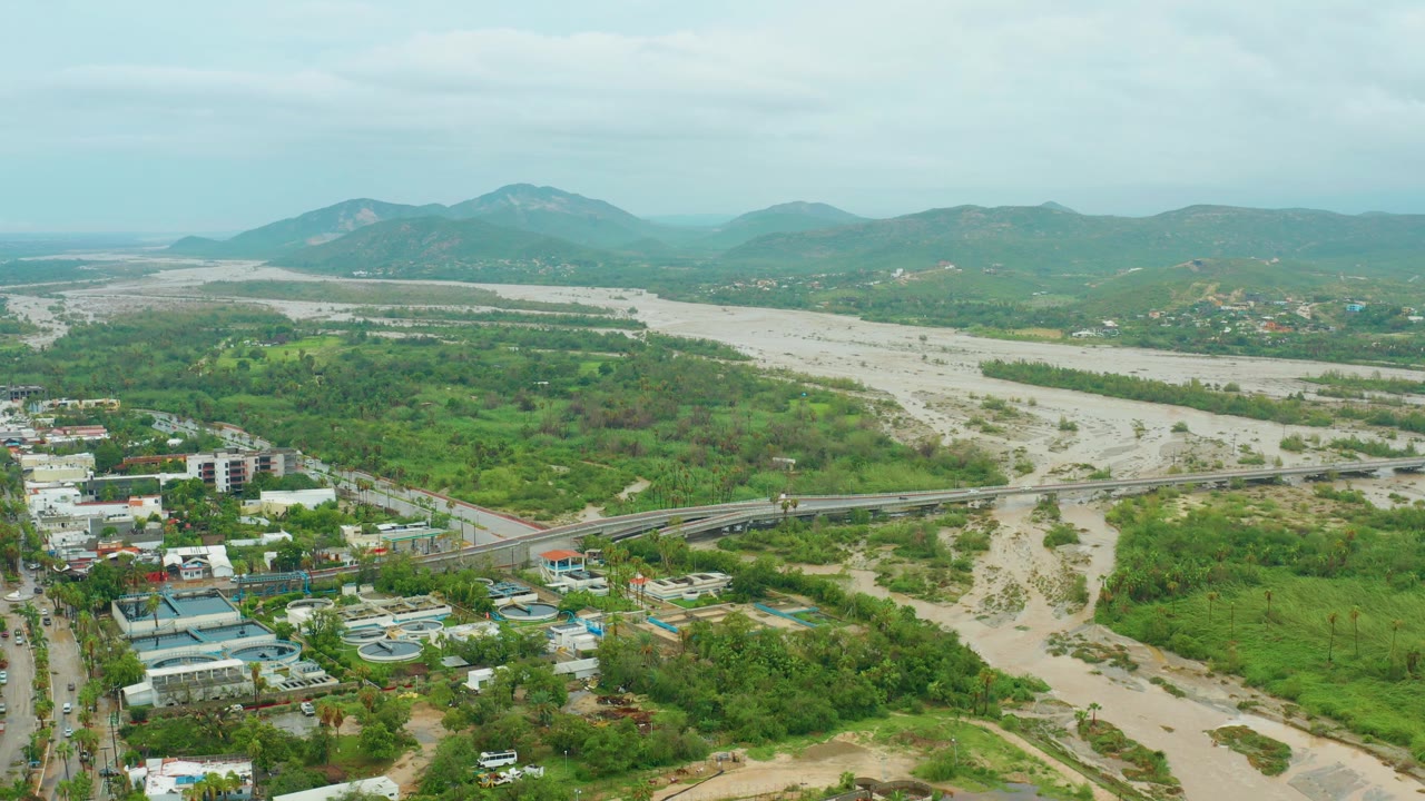 Mid-clip aerial view in San Jose del Cabo with pale floodwater and exposed sandy riverbank, a bridge crossing the lower frame, clusters of buildings beside green vegetation, distant hills, and no visible people.