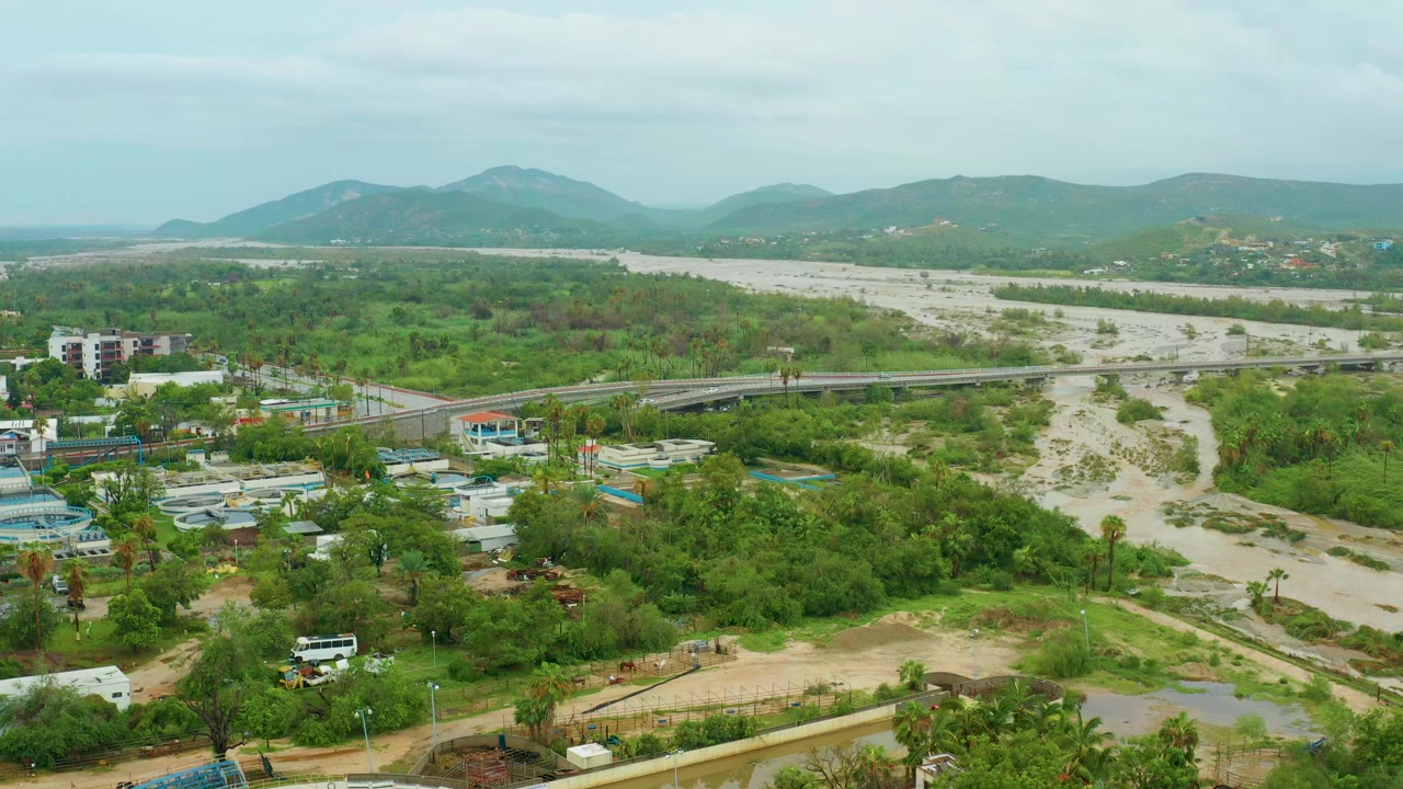 At 0:00, a high aerial view over San Jose Del Cabo, Baja California Sur shows a muddy flooded river beside green vegetation, a bridge, foreground buildings, distant hills, and no visible people.