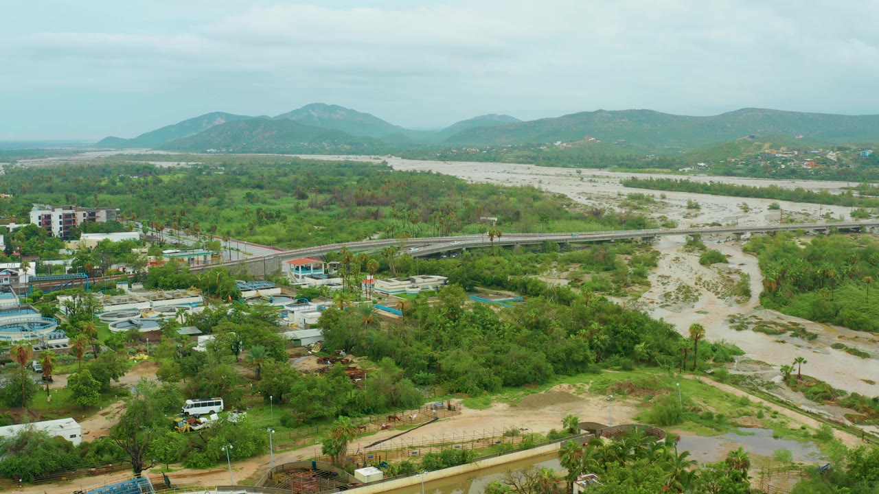 At 0:02, a high view in San Jose Del Cabo shows the pale muddy river channel stretching toward distant hills, with the bridge, green shrubs, foreground buildings, and no visible people.