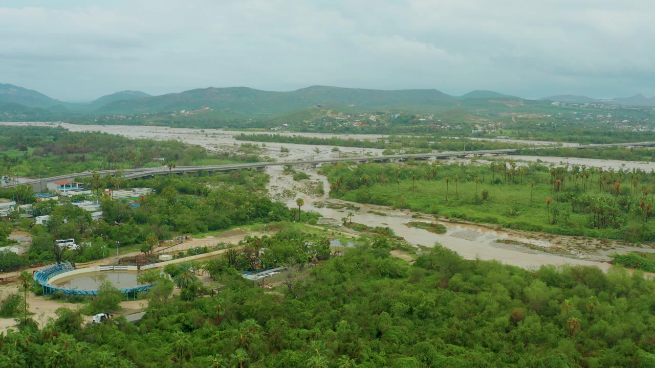 Wide aerial frame from San Jose del Cabo BCS Mexico showing a muddy river across green vegetation, a bridge in the midground, distant hills, overcast sky, and no visible people.