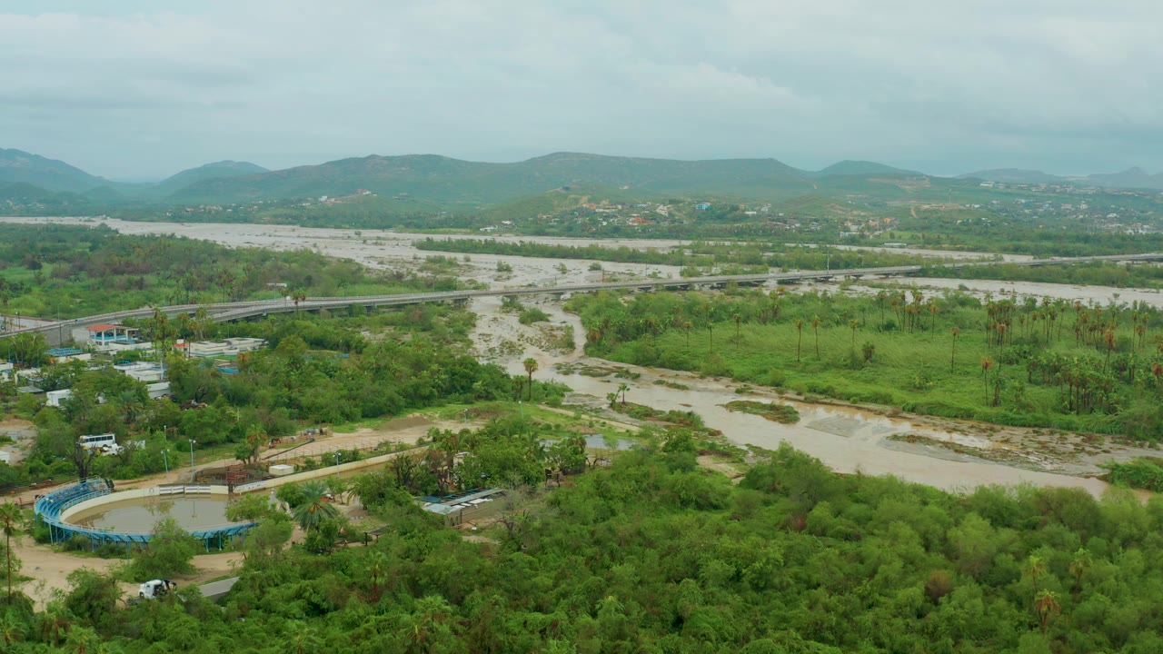 San Jose del Cabo BCS Mexico aerial view showing a broad muddy river bed, green valley vegetation, a bridge line across the scene, distant hills, cloudy light, and no visible people.