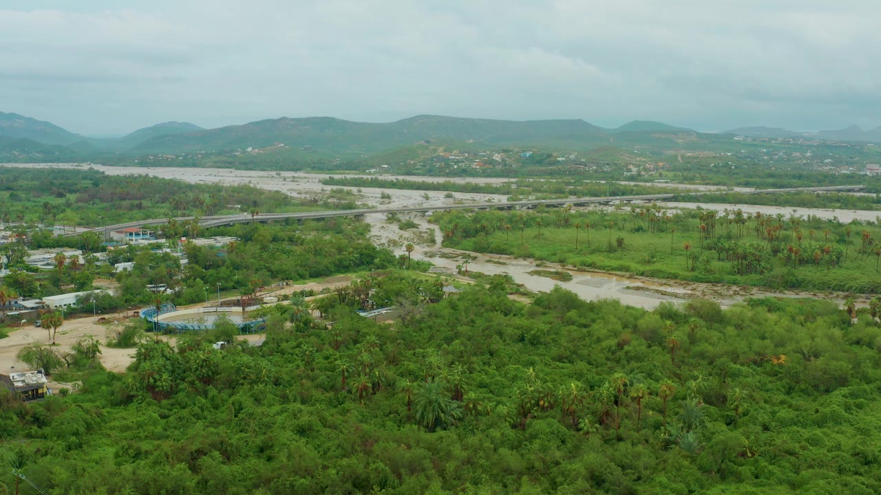 Wide aerial landscape with green trees, winding pale riverbed, scattered structures, roads, and hills.