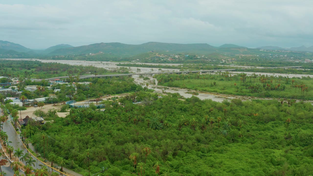 Aerial view over a lush green valley in Baja California Sur, Mexico, with a pale winding riverbed, scattered roads, distant hills, overcast sky, and no visible people.