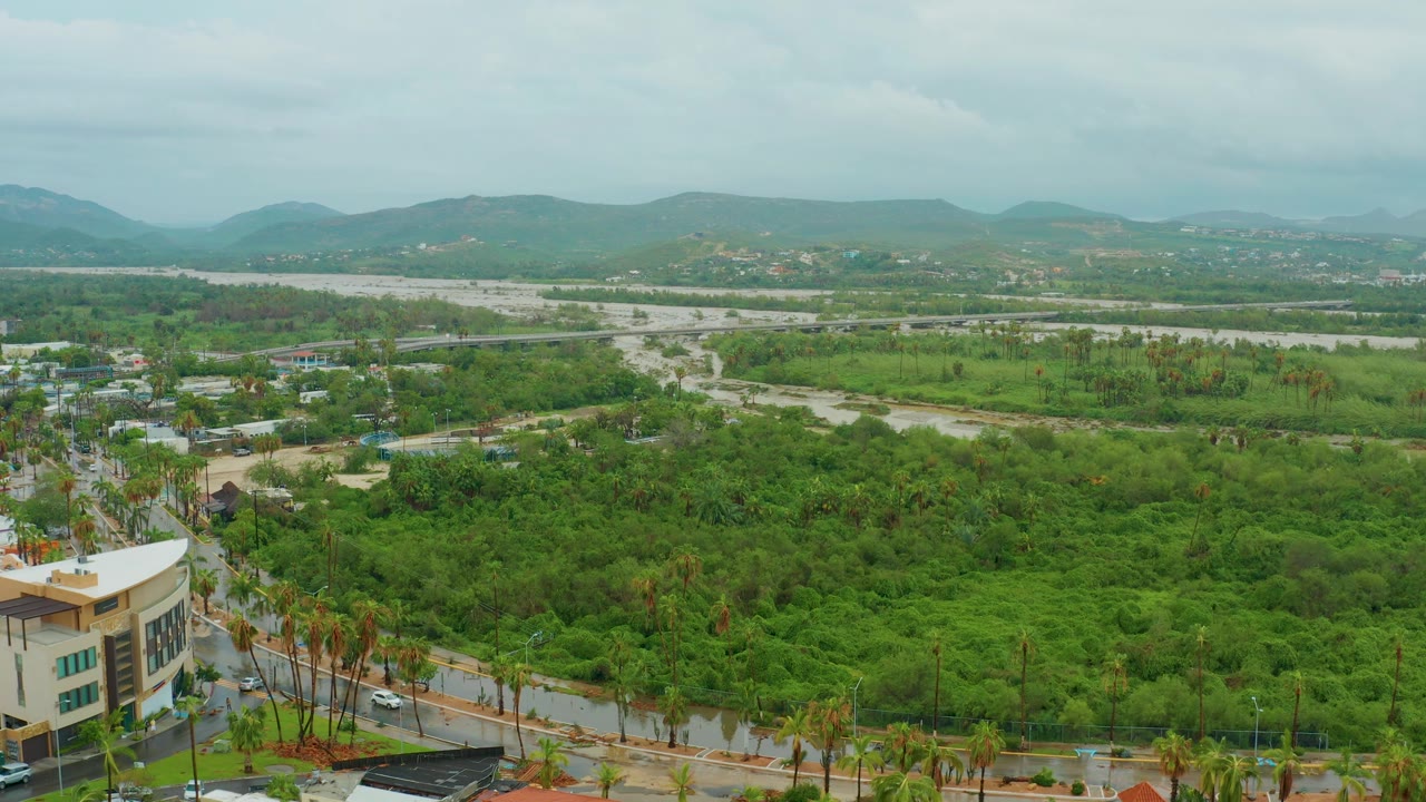 Wide aerial landscape with a road through greenery, scattered buildings, riverbed, and hills.