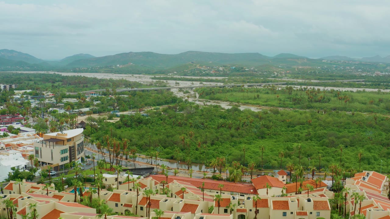 Aerial frame in San Jose del Cabo showing the residential rooftops shifted farther across the foreground, a light-colored multi-story building, a broad green corridor, distant hills, and no visible people.