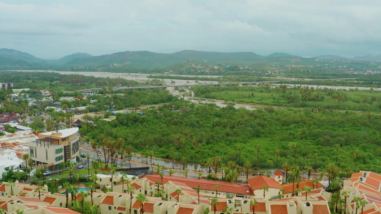 Aerial frame over San Jose del Cabo with red roofs along the lower edge, palm-lined streets, a large green area in the center, pale open ground near the horizon, mountains, and no visible people.