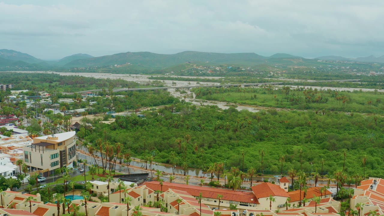 Aerial neighborhood view with roads, greenery, rooftops, and distant hills.