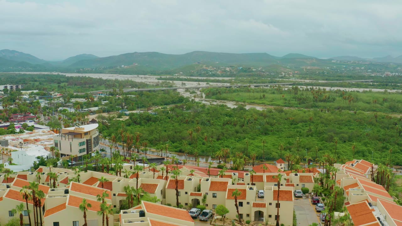 Opening aerial frame of San Jose del Cabo, Baja California Sur, Mexico, with red-tiled homes along the bottom edge, palms and a pale building at left, broad green fields, distant hills, and no visible people.