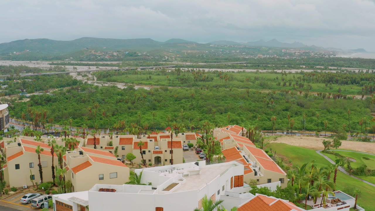 Early aerial frame over San Jose del Cabo residential buildings with terracotta roofs in the foreground, broad green land beyond, distant hills, overcast sky, and no visible people.