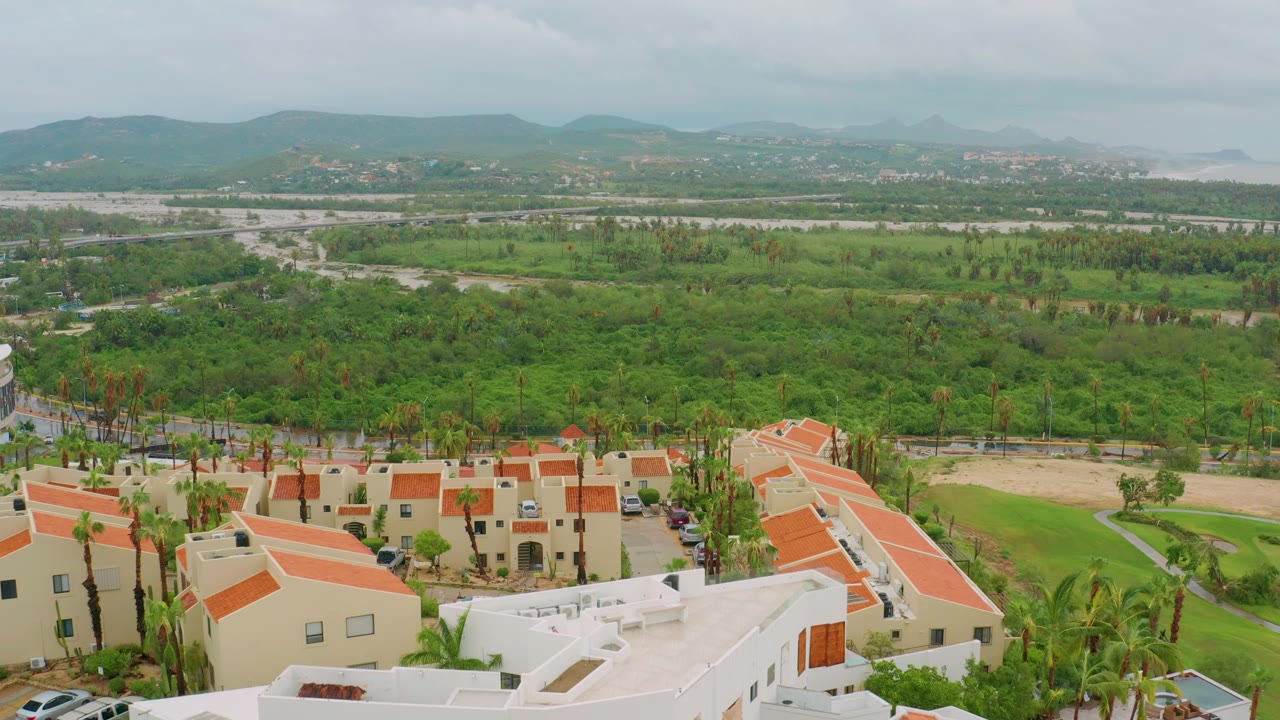 Mid-clip aerial view in San Jose del Cabo with orange terracotta roofs and white buildings at the bottom, green fields and palms across the center, mountains in the distance, and no visible people.