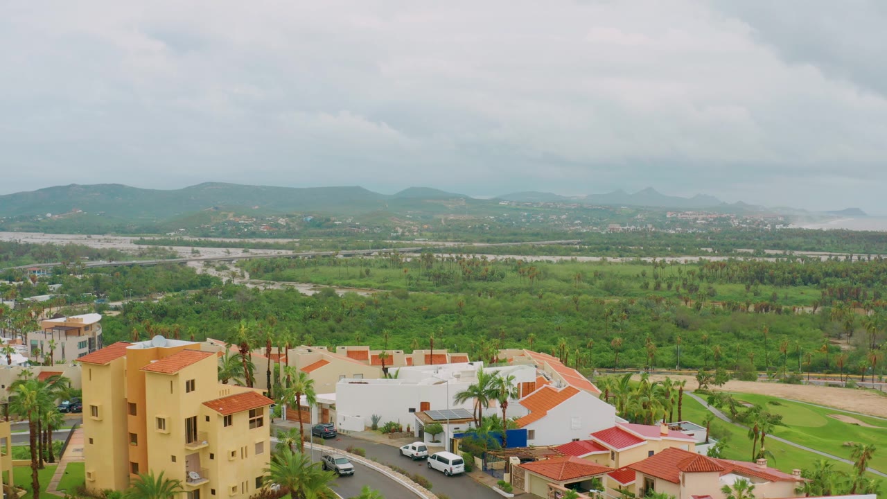 Opening aerial frame over San Jose del Cabo homes with red tile roofs, palms, a green valley, distant hills, cloudy sky, and no visible people.