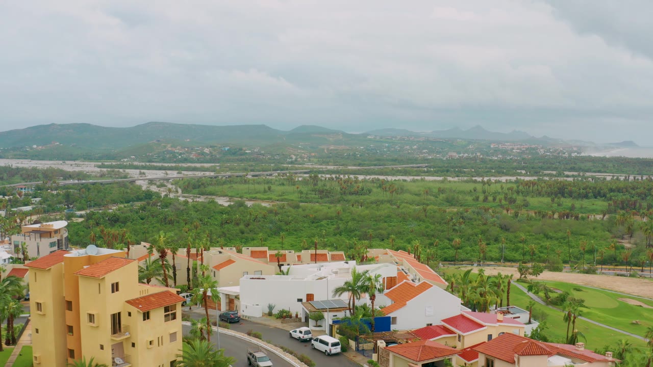San Jose del Cabo residential blocks with beige and white buildings, red tile roofs, palms, a road with vehicles, green fields, distant hills, and no visible people.