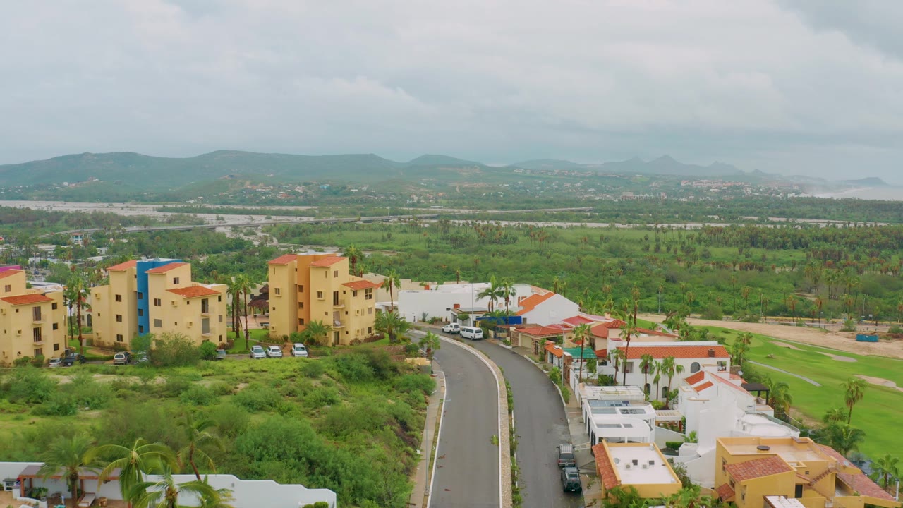 Opening aerial frame over San Jose Del Cabo showing yellow residential buildings, white homes with red roofs, a curving road, green lots, distant hills, cloudy sky, and no visible people.