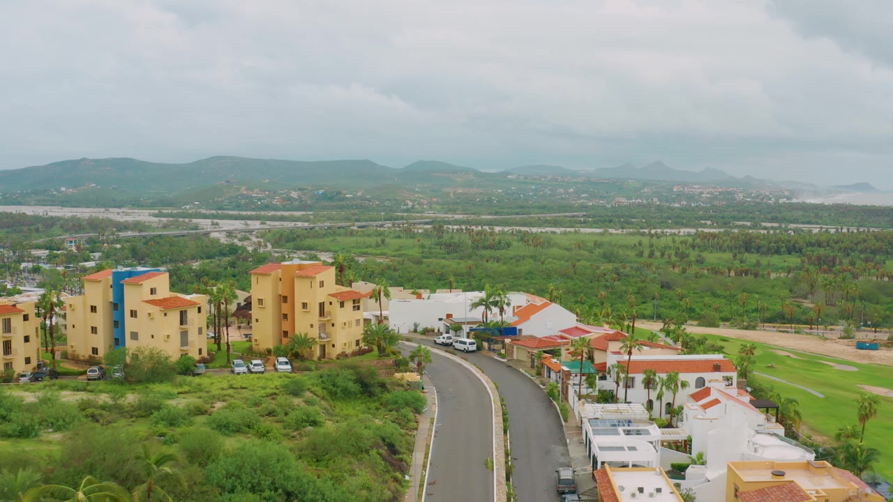 San Jose Del Cabo frame showing parked cars near yellow residential blocks, red-roofed white homes along the road, broad green midground, distant mountains, overcast light, and no visible people.