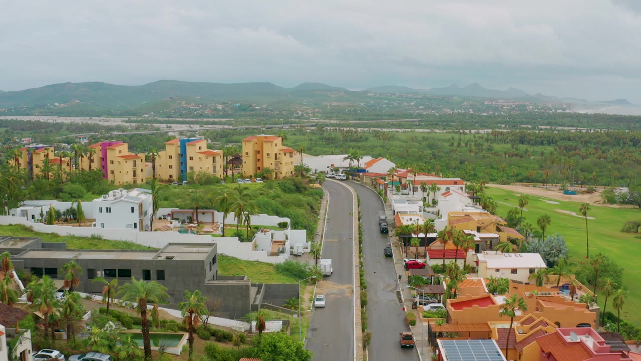 Aerial view in Baja California Sur, Mexico, showing a curved residential road, white homes, colorful apartment-style buildings, green lots, distant mountains, cloudy sky, and no visible people.