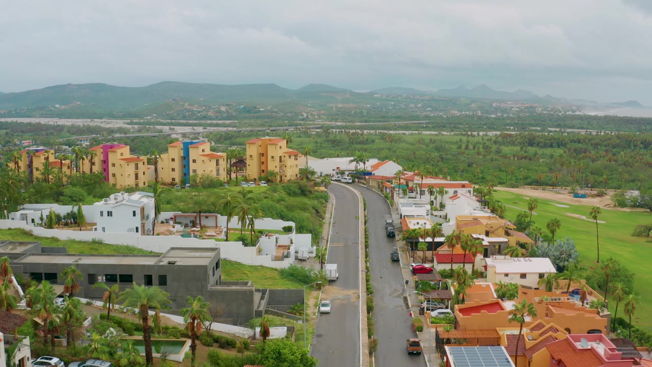Elevated frame in Baja California Sur, Mexico, with a white van on the curved road, low white homes, palm trees, colorful residential buildings, hazy mountains, cloudy sky, and no visible people.