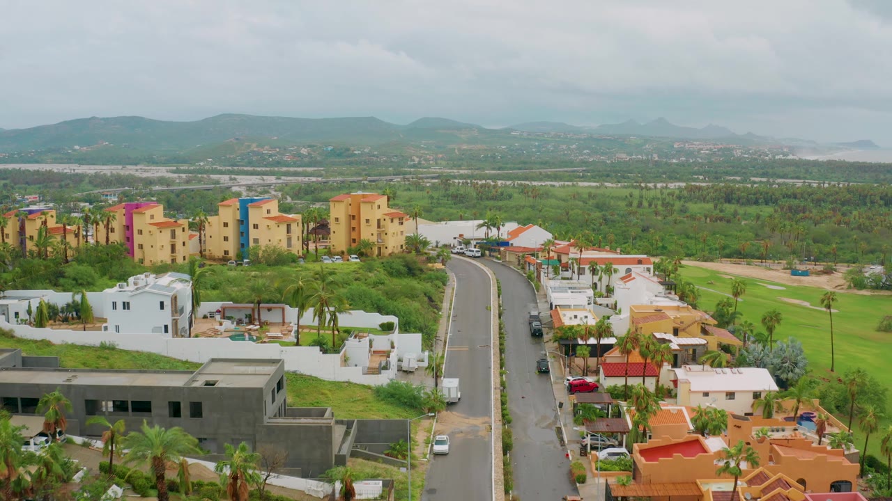 Aerial frame in Baja California Sur, Mexico, looking over a curving road, white walled homes, orange roofs, green open areas, colorful multi-story buildings, mountains, cloud cover, and no visible people.
