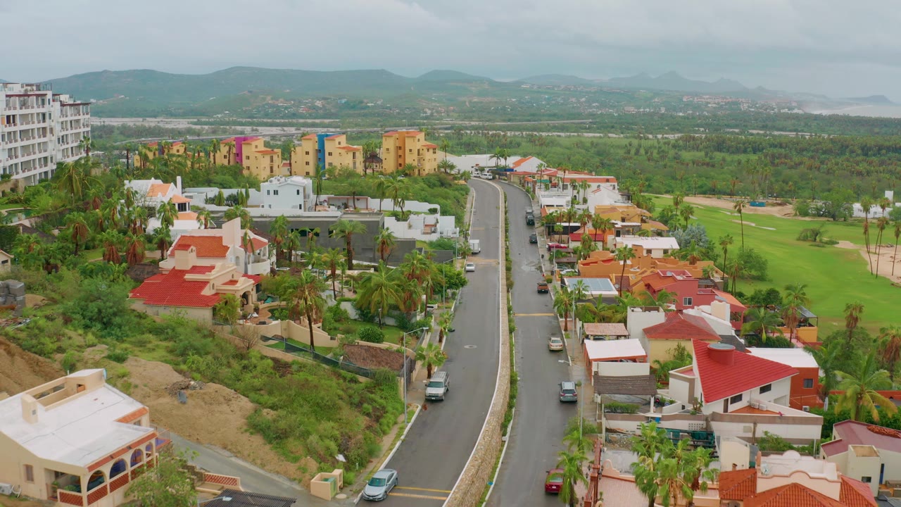 Elevated view in San Jose Del Cabo, Baja California Sur, looking over a curved residential road, red-roof houses, palms, green lots, distant hills, and no visible people.