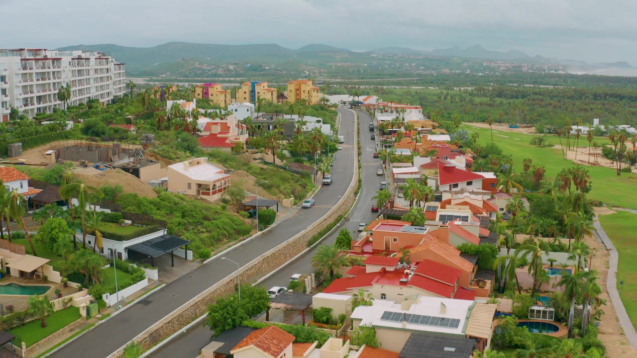 Aerial frame from San Jose del Cabo showing a curving road, red-roofed homes, palms, golf-course grass, a white mid-rise at left, distant hills, and no visible people.