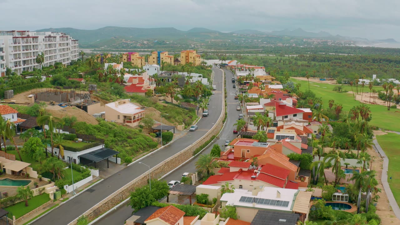 San Jose del Cabo aerial view with the road centered, red and white houses in the foreground, green golf-course fairways to the right, hazy hills beyond, and no visible people.