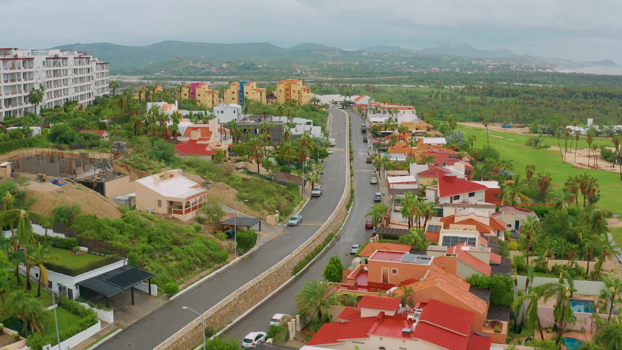 Aerial residential neighborhood with a winding road, construction site, green lawns, and distant hills.