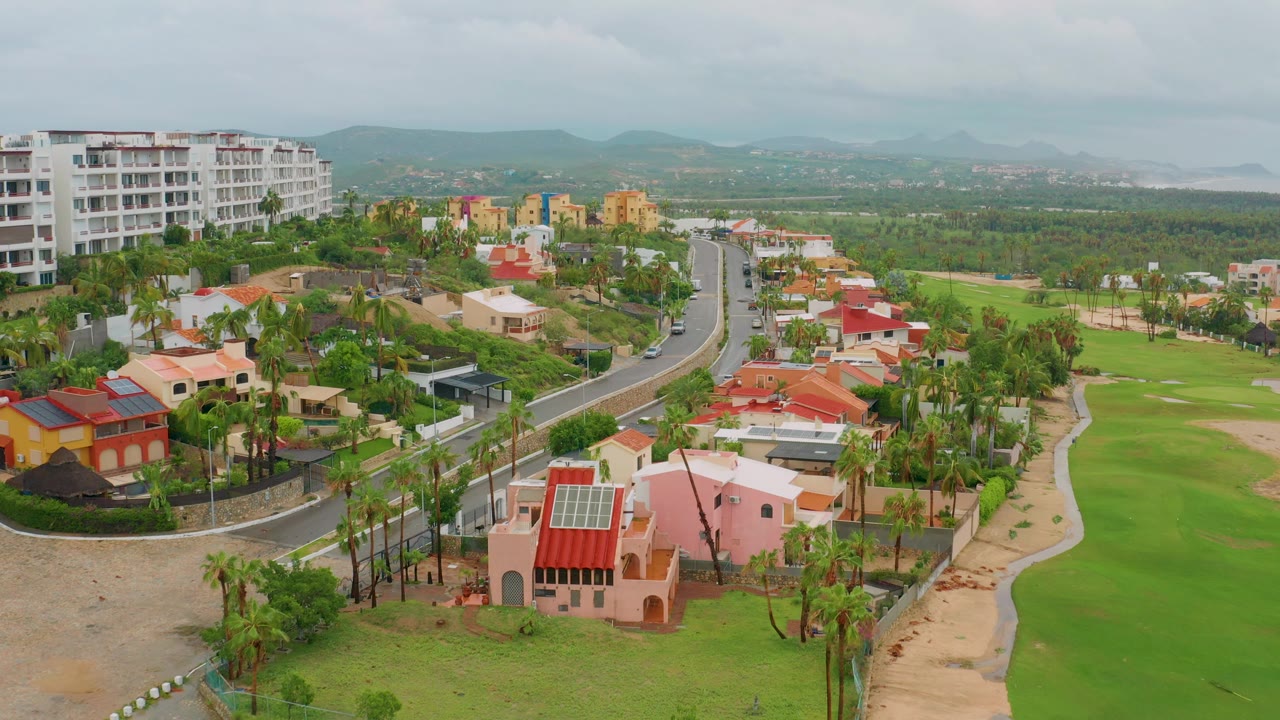 Aerial opening frame over San Jose del Cabo BCS Mexico showing red-roof and pastel homes, a curved road, palm trees, green golf-course fairway, distant hills, and no visible people.