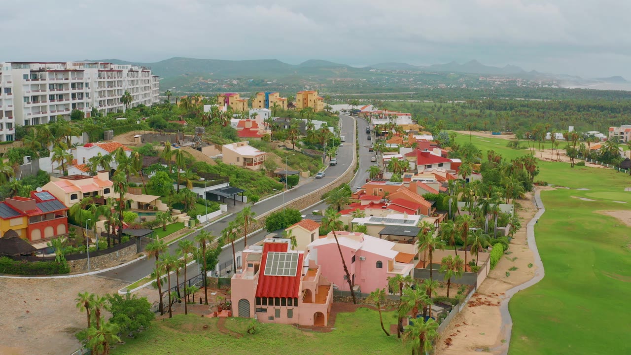 San Jose del Cabo BCS Mexico aerial frame showing the pink foreground house lower right, palm-lined homes along the curved road, golf-course grass to the side, distant mountains, and no visible people.