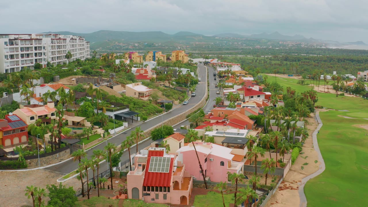 Aerial frame of a curving neighborhood road, homes, palm trees, golf grass, and distant hills.