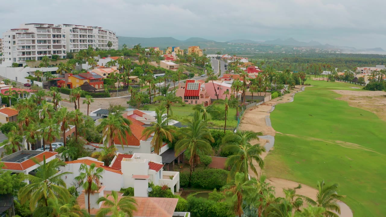 Aerial frame over San Jose del Cabo resort homes, palm trees, a green golf fairway, distant mountains, and an overcast sky, with no visible people.