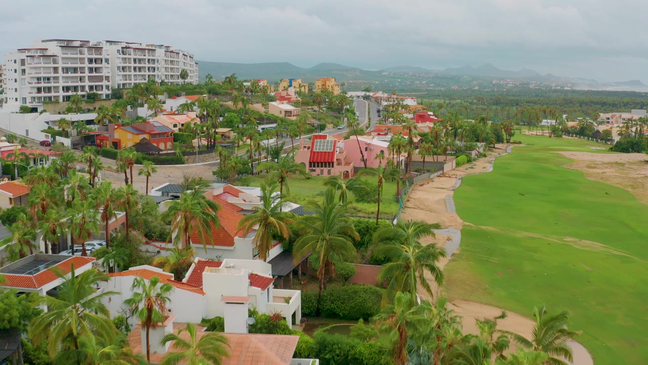 San Jose del Cabo aerial view with foreground white villas, tall palms, a curving road, the golf course to the right, and hazy mountains under cloud cover, with no visible people.