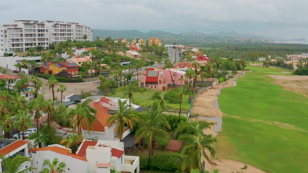 Wide San Jose del Cabo aerial with a large white apartment building at upper left, clustered resort homes, palm-lined roads, a green fairway, and distant mountains, with no visible people.