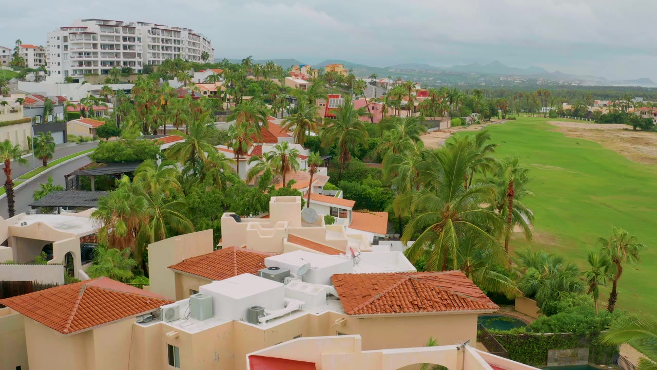 Aerial view over San Jose Del Cabo homes with red tile roofs in the foreground, palms around the neighborhood, a green field to the right, a white multi-story building behind, and no visible people.