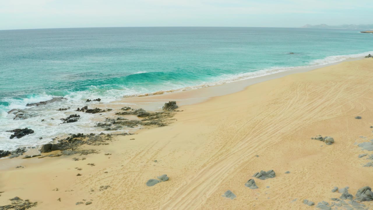 Elevated view of Laguna Beach Los Cabos with turquoise water, white surf, pale sand, scattered rocks, tire tracks, hazy horizon, and no visible people.