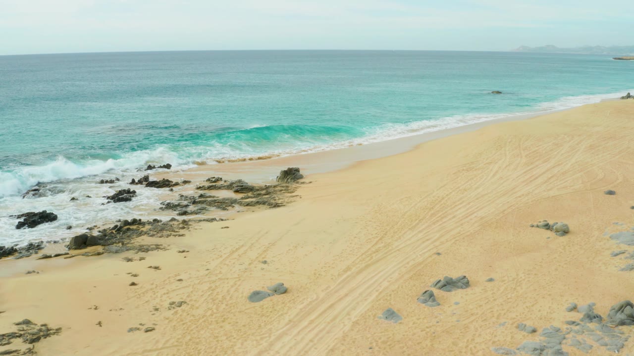 Wide aerial frame over Laguna Beach Los Cabos showing a clean band of turquoise water, surf foam near the rocks, open sand, tire tracks, and no visible people.
