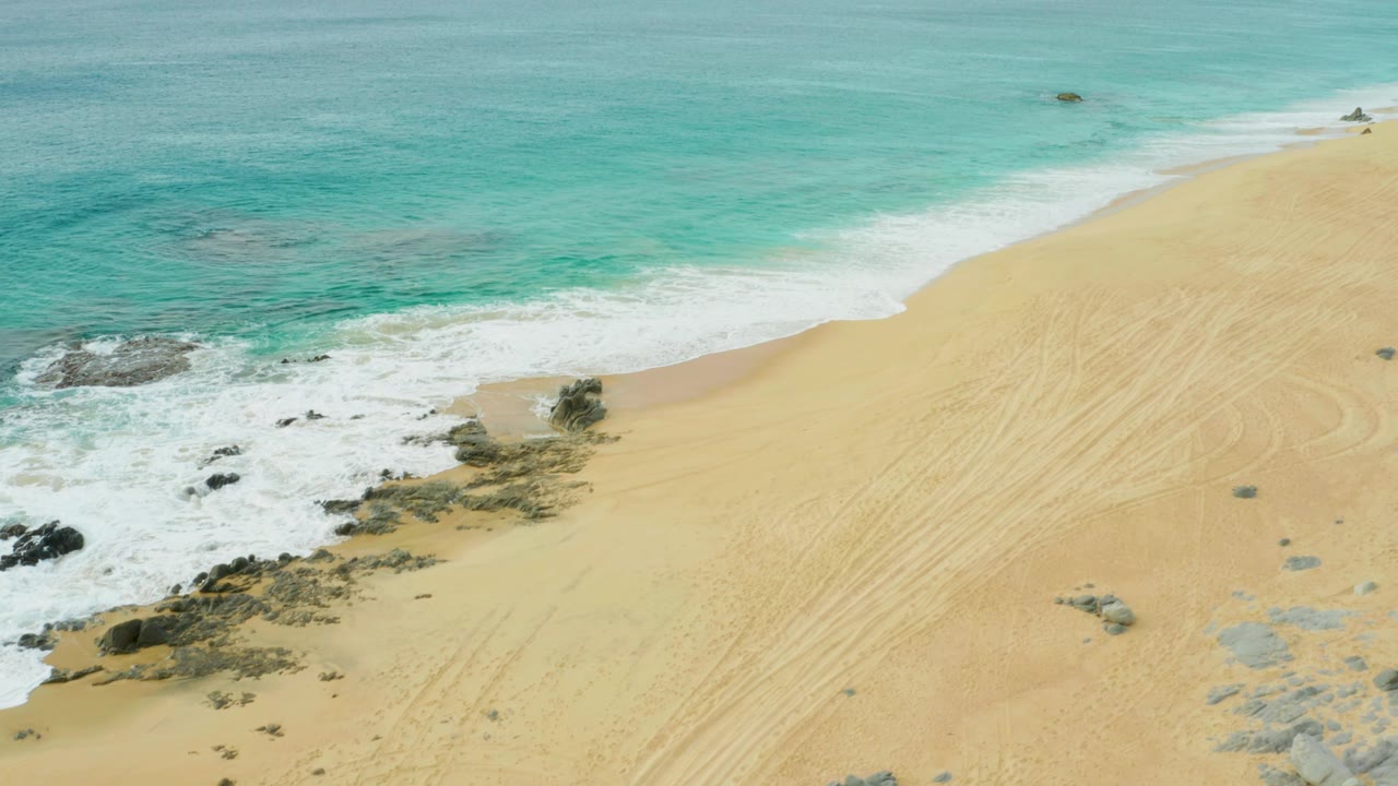 Aerial view of Laguna Beach Los Cabos with foamy surf pushing farther across the rocky shoreline and a broad pale sand area to the right, with no visible people.
