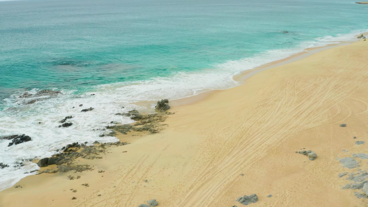 High aerial view at Laguna Beach Los Cabos where white surf wraps around low rocks on the left and pale sand with faint tracks fills the right side, with no visible people.