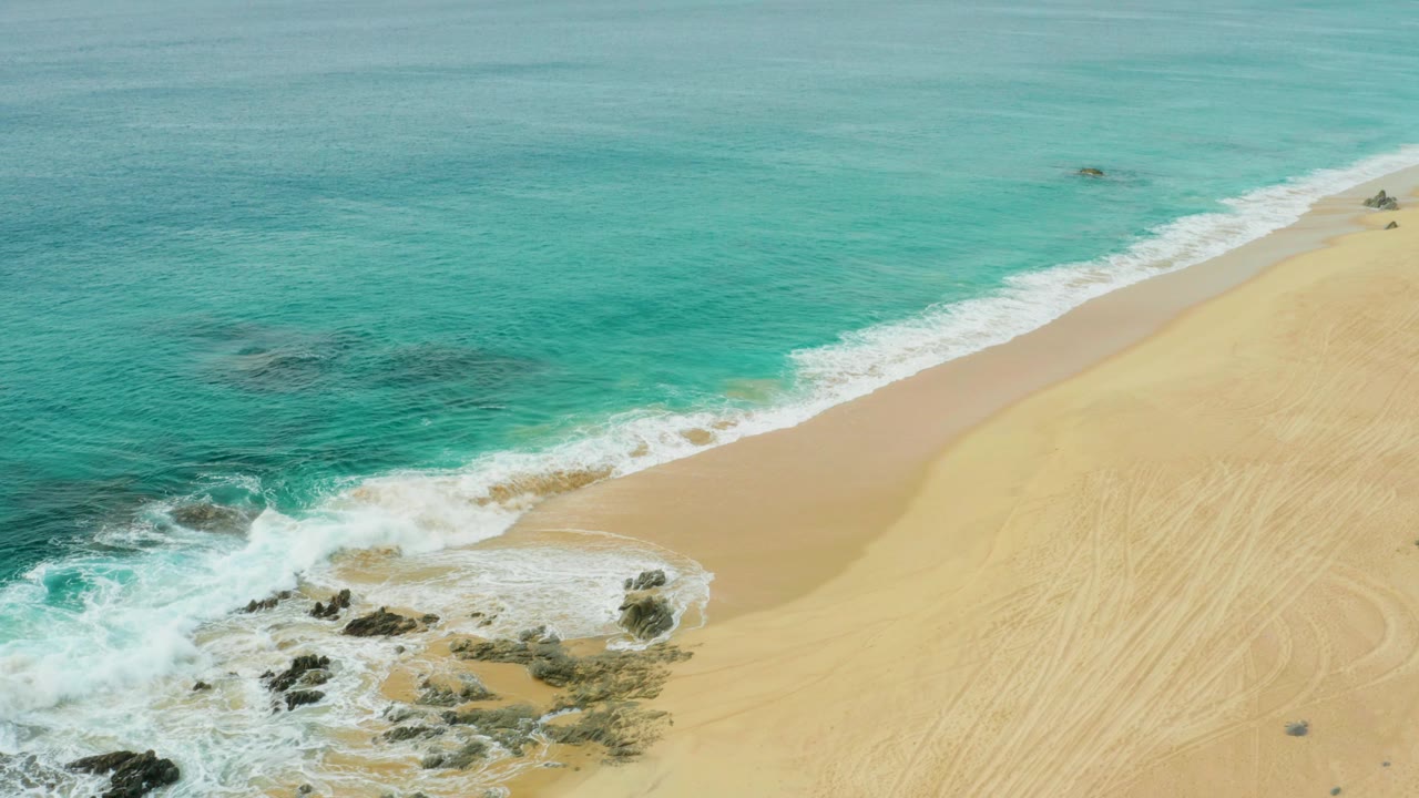 Aerial view at Laguna Beach Los Cabos, Mexico, with turquoise water, white surf, pale sand, scattered rocks near the lower edge, and no visible people.