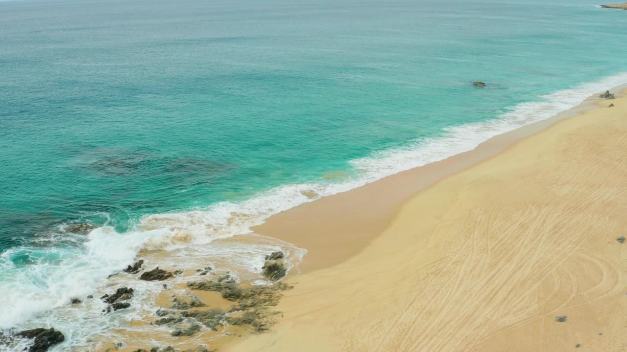 Aerial view at Laguna Beach Los Cabos, Mexico, with a wider band of turquoise water, a diagonal white surf edge, exposed rocks near the lower left, marked sand on the right, and no visible people.