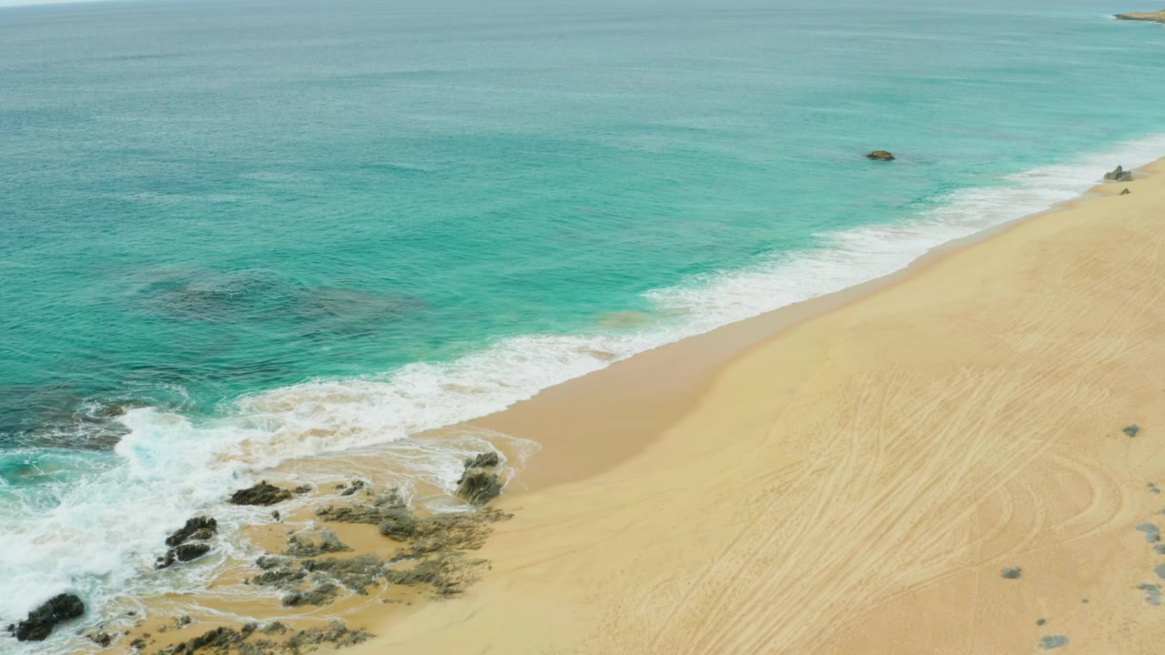 Aerial beach view with surf, rocks, and pale sand.